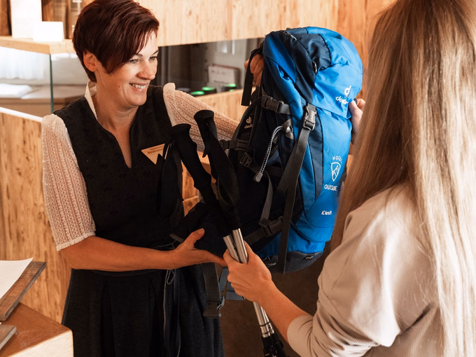 Mitarbeiterin Karin übergibt einem Gast einen blauen Wanderrucksack und Teleskopstöcke am Empfang Naturhotel Outside in Matrei in Osttirol