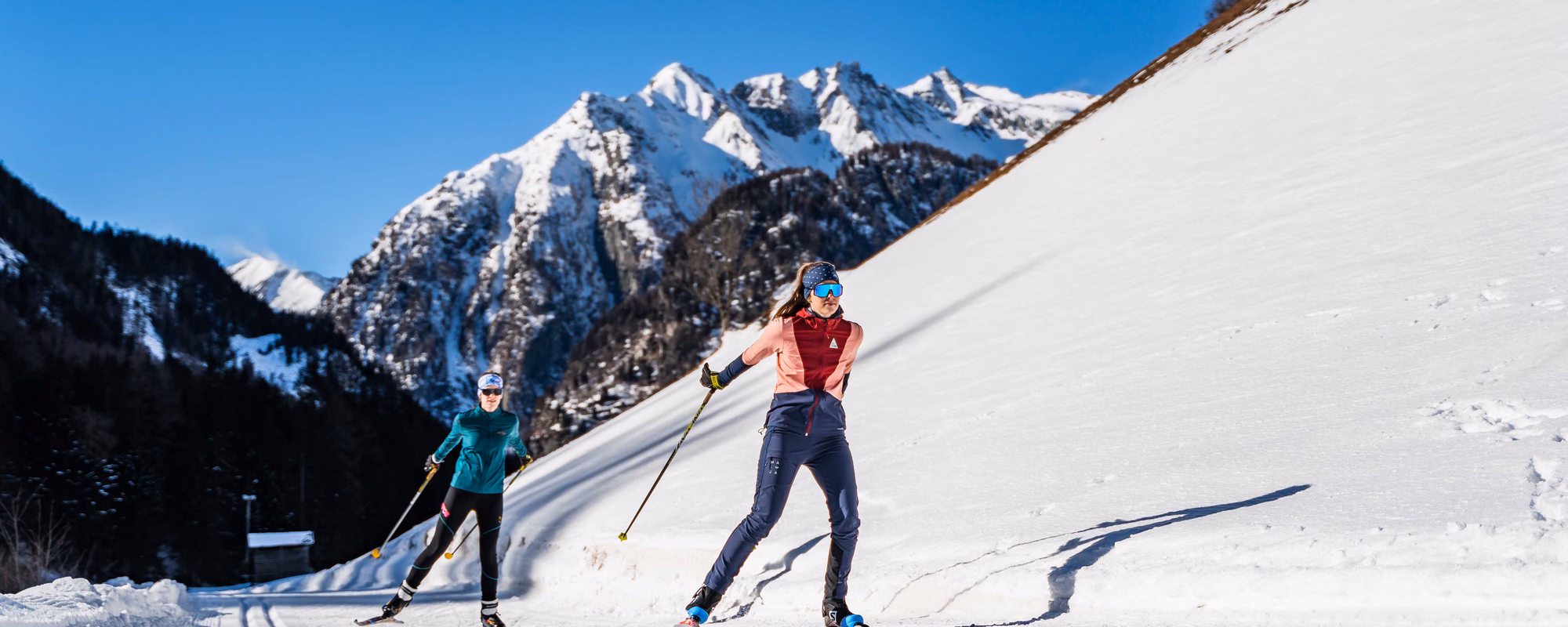 Zwei Langläuferinnen auf der breiten Skating-Loipe in Prägraten vor schroffen, schneebedeckten Berggipfeln.