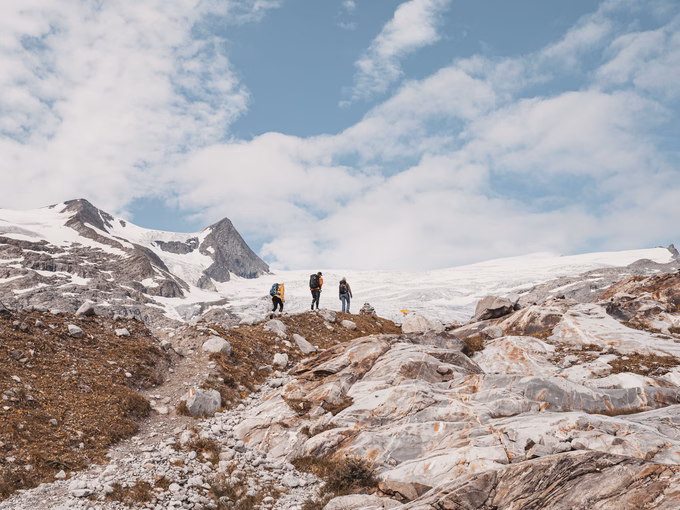 Drei Wanderer mit Rucksäcken auf einem alpinen Steinpfad vor dem Schlatenkees-Gletscher und schneebedeckten Bergen.