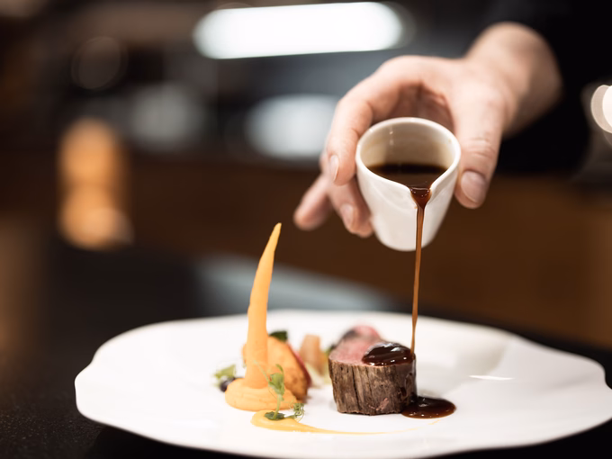 Ein Koch verfeinert ein rosa gebratenes Rindsfilet im Speckmantel mit dunkler Sauce auf einem weißen Porzellanteller im Naturhotel Outside in Matrei in Osttirol.