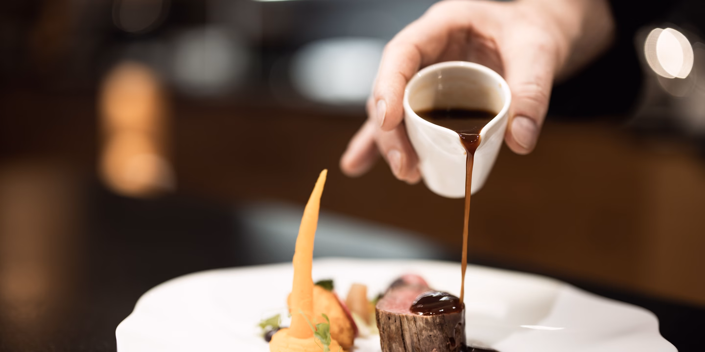 Ein Koch verfeinert ein rosa gebratenes Rindsfilet im Speckmantel mit dunkler Sauce auf einem weißen Porzellanteller im Naturhotel Outside in Matrei in Osttirol.