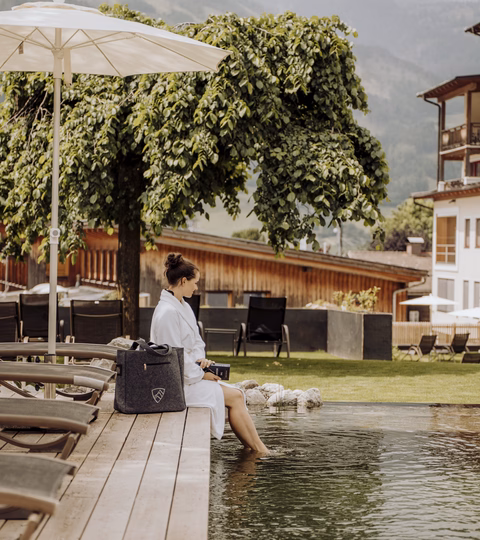 Frau im weißen Bademantel sitzt am Holzsteg des Naturbadeteichs beim Naturhotel Outside in Matrei in Osttirol.