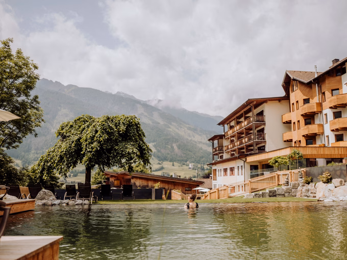 Frau schwimmt im chlorfreien Naturpool mit Blick auf das Hotelgebäude und die Bergkulisse beim Naturhotel Outside in Matrei in Osttirol.