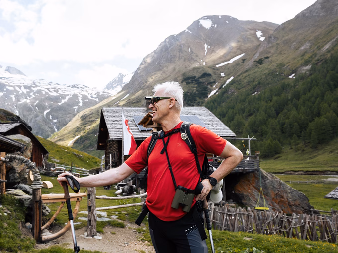 Seniorchef Sepp vor den traditionellen Holzhütten der Steineralm mit Gebirgspanorama nahe dem Naturhotel Outside in Matrei in Osttirol.