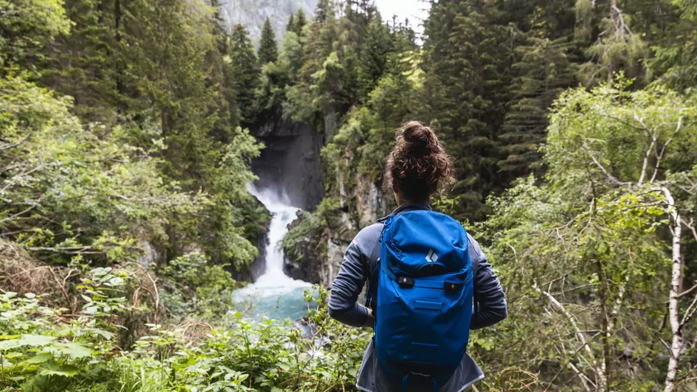 Frau mit Rucksack steht vor kleinem Wasserfall inmitten unberührter Natur
