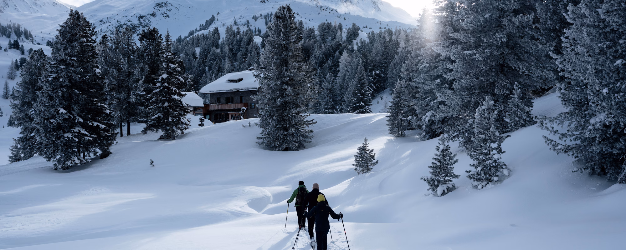 Drei Schneeschuhwanderer folgen einer Spur im tiefen Schnee in Richtung einer verschneiten Berghütte, Naturhotel Outside in Matrei in Osttirol.