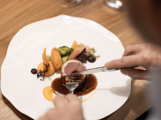 Ein Gast schneidet ein rosa gebratenes Rindsfilet im Speckmantel mit Beilagen und Rotwein an einem Holztisch im Naturhotel Outside in Matrei in Osttirol.