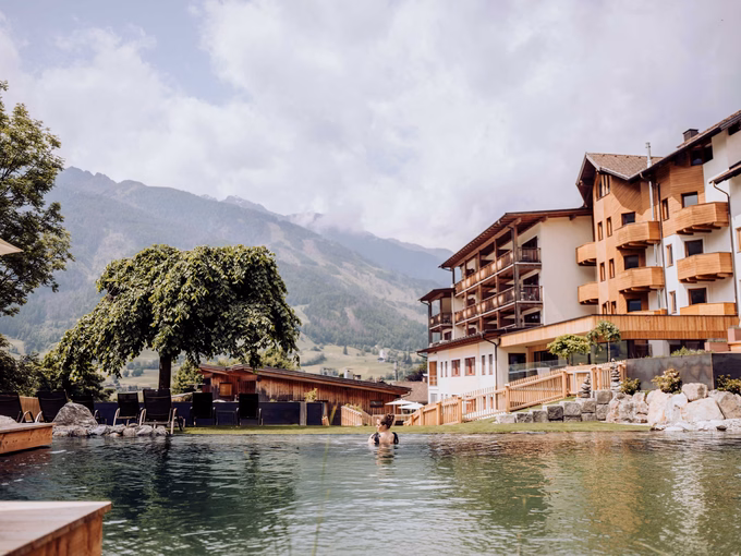 Badegast im natürlichen Badeteich mit Blick auf das Hotelgebäude und die Berge beim Naturhotel Outside in Matrei in Osttirol