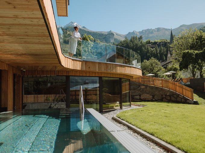 Beheizter Außenpool und moderne Holzarchitektur mit einer Frau auf dem Balkon beim Naturhotel Outside in Matrei in Osttirol.