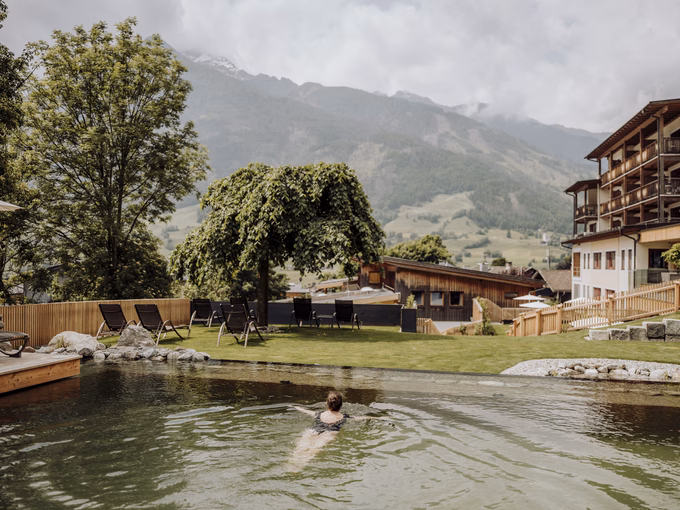 Panoramaaufnahme der Gartenanlage mit schwimmender Person im Naturbadeteich beim Naturhotel Outside in Matrei in Osttirol.
